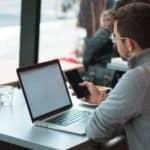 man sitting near table with laptop and smartphone near window