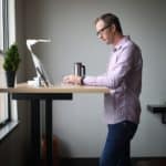 man in pink dress shirt and blue denim jeans standing beside brown wooden table